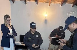 Una mujer en blazer oscuro observa a tres policías organizando evidencias en una mesa cubierta de objetos como computadoras y teléfonos.