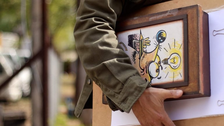 Persona con brazos visibles sostiene un marco con dibujo de bombilla en un entorno desenfocado y al aire libre.