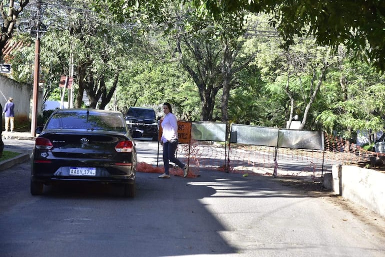 Mujer con camisa clara y jeans oscuros camina frente a auto negro, en calle bloqueada por cercas anaranjadas.