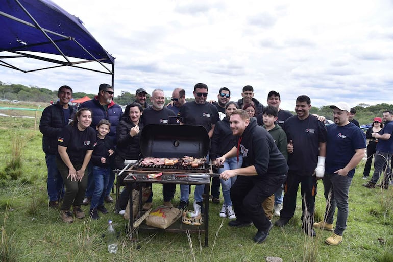 Diversos participantes sonríen y se agrupan alrededor de un asador. Hombre con sudadera negra cocina carne en un ambiente festivo.