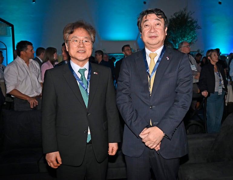 Dos hombres en trajes oscuro y azul, con corbatas verde y amarilla, en evento social iluminado con luces azules.