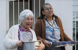 Mujeres sonrientes en exterior. Una sostiene olla metálica, la otra lleva delantal floral y suéter marrón.