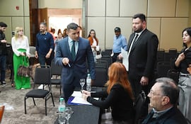 Hombre con traje y corbata dialoga con mujer de cabello rojo en mesa con documentos, rodeados de asistentes en salón de eventos.