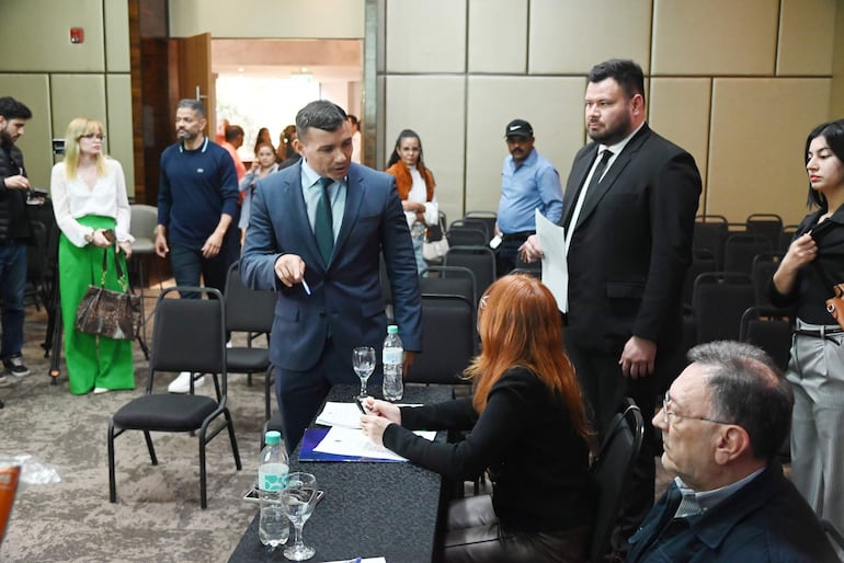 Hombre con traje y corbata dialoga con mujer de cabello rojo en mesa con documentos, rodeados de asistentes en salón de eventos.