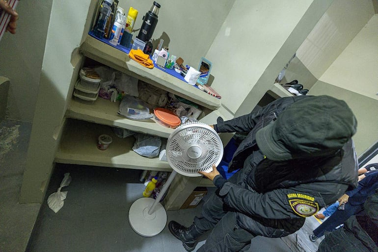 Hombre con chaleco oscuro y gorro, inspecciona ventilador en ambiente desordenado, rodeado de objetos y otros hombres.