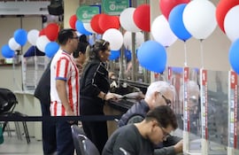 Mujer de pie en mostrador asistiendo a hombre canoso, mientras otro hombre joven revisa documentos en un ambiente decorado con globos.