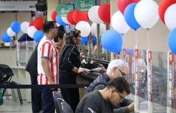 Mujer de pie en mostrador asistiendo a hombre canoso, mientras otro hombre joven revisa documentos en un ambiente decorado con globos.