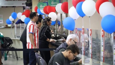 Mujer de pie en mostrador asistiendo a hombre canoso, mientras otro hombre joven revisa documentos en un ambiente decorado con globos.