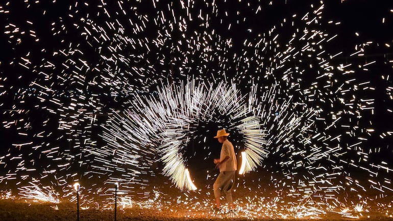 Persona vestida con atuendo claro y sombrero, danzando entre chispas brillantes en un ambiente nocturno.
