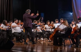 Luis Álvarez dirigiendo a la Orquesta Nacional de Música Popular, músicos concentrados en sus instrumentos de cuerda en escenario formal.