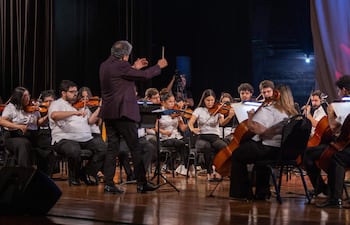 Luis Álvarez dirigiendo a la Orquesta Nacional de Música Popular, músicos concentrados en sus instrumentos de cuerda en escenario formal.