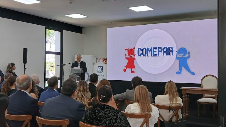 Orador canoso en traje oscuro en podio de cristal con audiencia formal, mujer a su lado observa atentamente.