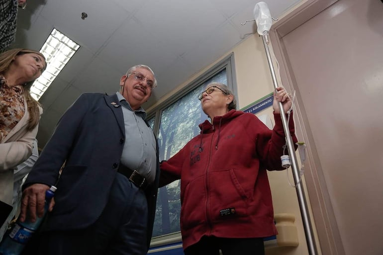Isaías Fretes en blazer interactúa con mujer en abrigo rojo sosteniendo un soporte para suero, en un entorno hospitalario.