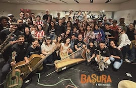 Grupo de músicos sonrientes sosteniendo instrumentos en un ambiente festivo, con carteles del musical 'Basura' de fondo.