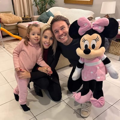 Niña en mono rosa sostiene la mano de una mujer en abrigo negro, mientras un hombre con camiseta gris muestra un peluche de Minnie Mouse.