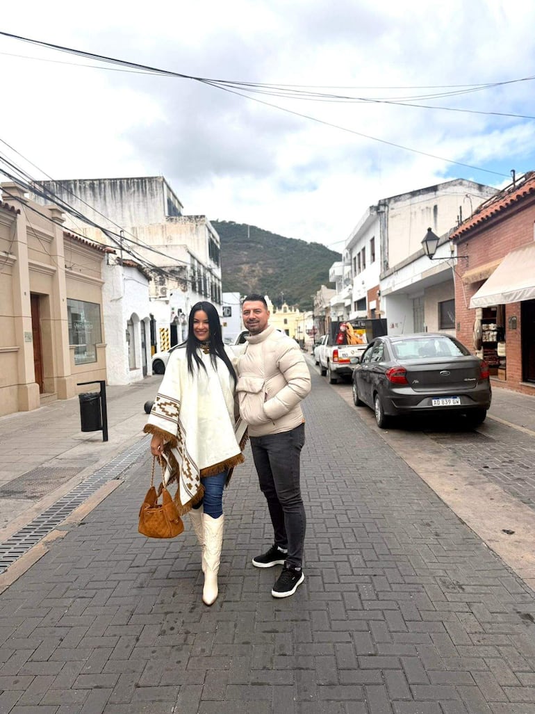 Gloria Vera con poncho claro y botas blancas, junto a Julio Cáceres en abrigo claro, ambos sonrientes en una calle adoquinada.