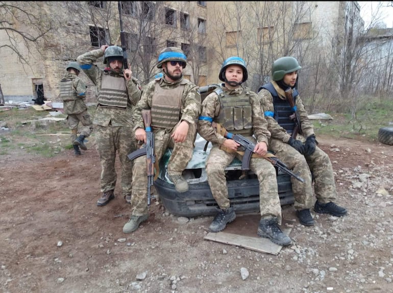 Seis soldados en uniformes de camuflaje y chalecos antibalas, dos de ellos de pie junto a un vehículo dañado en un paisaje desolado.