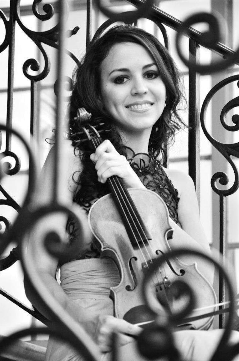 María Arrúa sonriente en vestido elegante sostiene un violín, sentada en escalera decorativa con fondo arquitectónico.