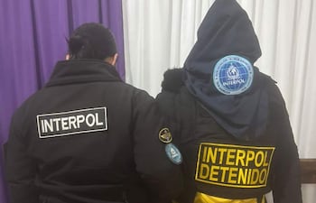 Dos agentes de Interpol con chaquetas negras y amarillas, en un entorno formal, durante un procedimiento de detención.