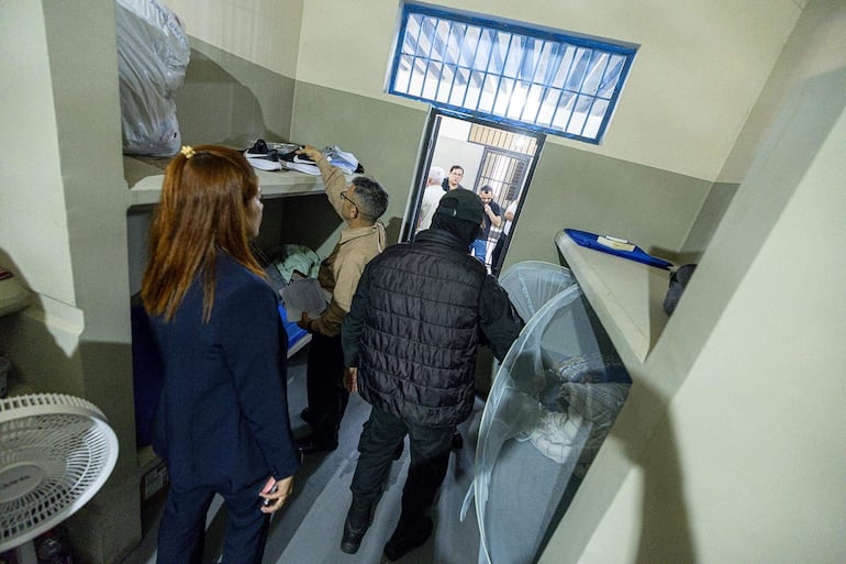 Mujer con cabello largo y rojo observa, mientras hombre con gafas señala con documento en mano, en celda penitenciaria con paredes claras.