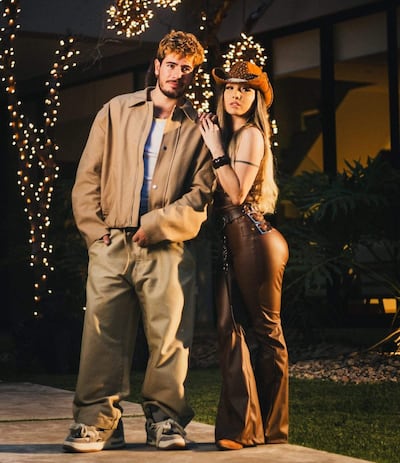Marilina Bogado con atuendo marrón y sombrero, junto a un hombre de chaqueta beige en un ambiente iluminado y festivo.