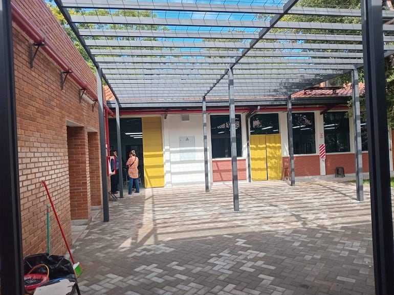 Dos personas, una con blusa marrón claro y pantalones oscuros, conversan en un patio con estructura metálica y paredes de ladrillo.