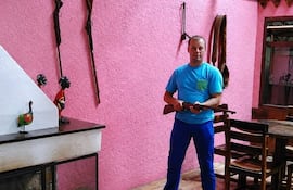 Hombre de pie en entorno rústico, sostiene un arma, viste camiseta azul y pantalones azules en ambiente con decoraciones de madera.
