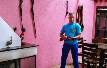 Hombre de pie en entorno rústico, sostiene un arma, viste camiseta azul y pantalones azules en ambiente con decoraciones de madera.