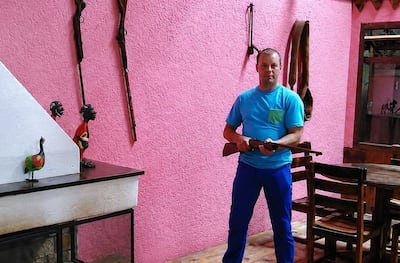 Hombre de pie en entorno rústico, sostiene un arma, viste camiseta azul y pantalones azules en ambiente con decoraciones de madera.