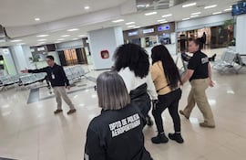 Grupo de cinco personas en el aeropuerto, tres mujeres con camisetas del Dpto. de Policía Aeroportuaria, hombre en abrigo negro hablando.