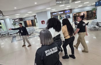 Grupo de cinco personas en el aeropuerto, tres mujeres con camisetas del Dpto. de Policía Aeroportuaria, hombre en abrigo negro hablando.