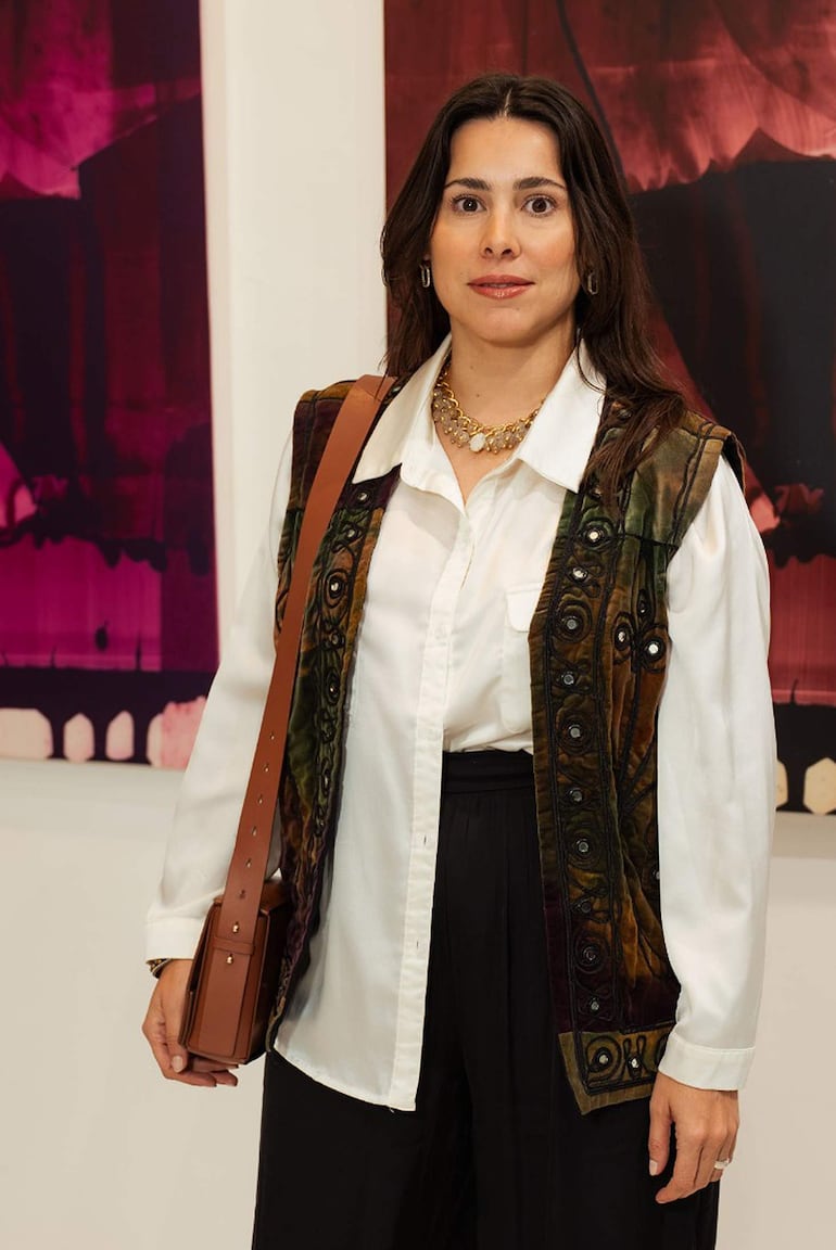 Mujer de pie en galería, viste chaleco multicolor y blusa blanca, sonriendo serenamente frente al arte abstracto de fondo.