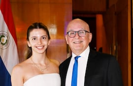 Mujer de cabello claro y blusa blanca sonríe junto a hombre en traje oscuro en evento con bandera paraguaya de fondo.