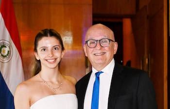 Mujer de cabello claro y blusa blanca sonríe junto a hombre en traje oscuro en evento con bandera paraguaya de fondo.