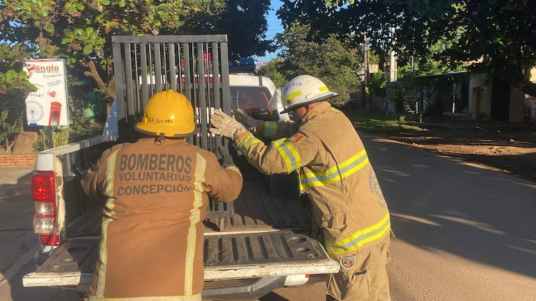 Bomberos de espaldas en uniforme marrón y cascos amarillo y blanco, cargando material en camioneta en una calle residencial.