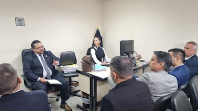 Un hombre en traje oscuro habla mientras Dalia López escucha atentamente. Otras personas observan en un ambiente serio y formal.