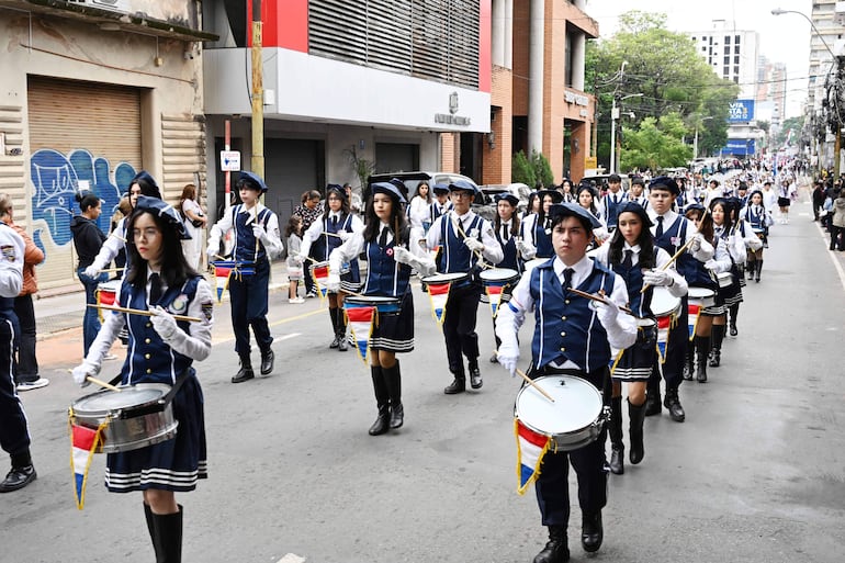Jóvenes en uniformes azul marino y blanco marchan, liderados por un joven gesticulando, observados por adultos y una niña.