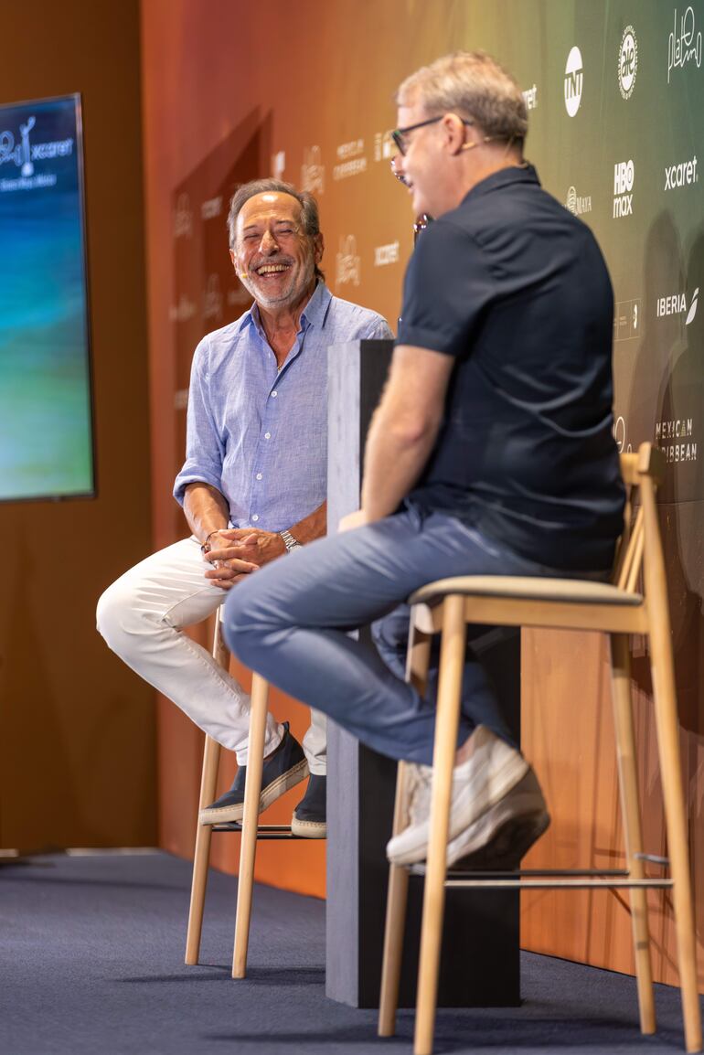 Francella con camisa azul claro y pantalones blancos sonríe mientras escucha a un hombre de camiseta oscura en un entorno colorido.
