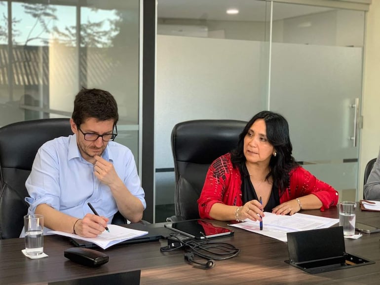 Emiliano Fernández escribe en un cuaderno mientras Paola Tapia lo observa, ambos en una oficina moderna y iluminada.