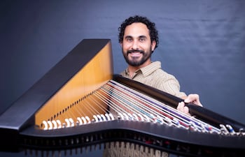 Hombre sonriente con cabello rizado y barba sostiene un arpa, vestido con camisa clara de rayas, en un entorno interior gris.