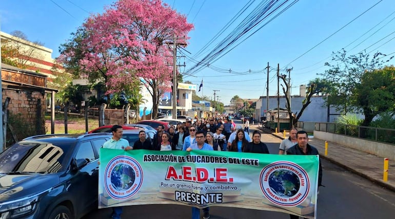 Participantes de la manifestación sostienen pancarta de la Asociación de Educadores del Este en una calle soleada, con expresión tranquila y felices.