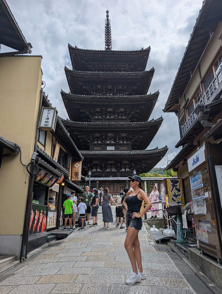 Mujer segura en camiseta y gorra, junto a turistas y una alta pagoda japonesa bajo un cielo nublado.