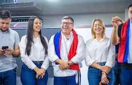 Hombre con bandera roja, blanca y azul sonríe entre mujeres de camisa blanca. Ambiente festivo interior con expresiones alegres.