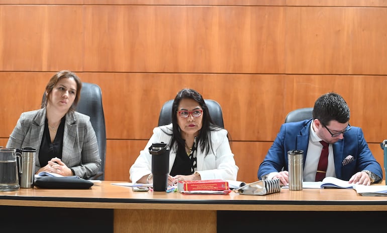 Tania Irún en chaqueta a cuadros, otra mujer con abrigo blanco y un hombre en traje azul revisan documentos en ambiente formal.