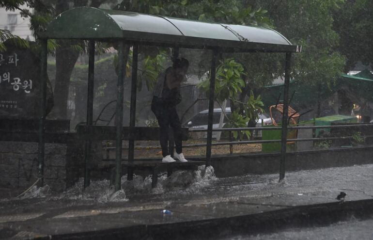 Una pasajera se resguarda precariamente de la lluvia parada en el banco de una parada de colectivos, mientras el raudal corre calle abajo.