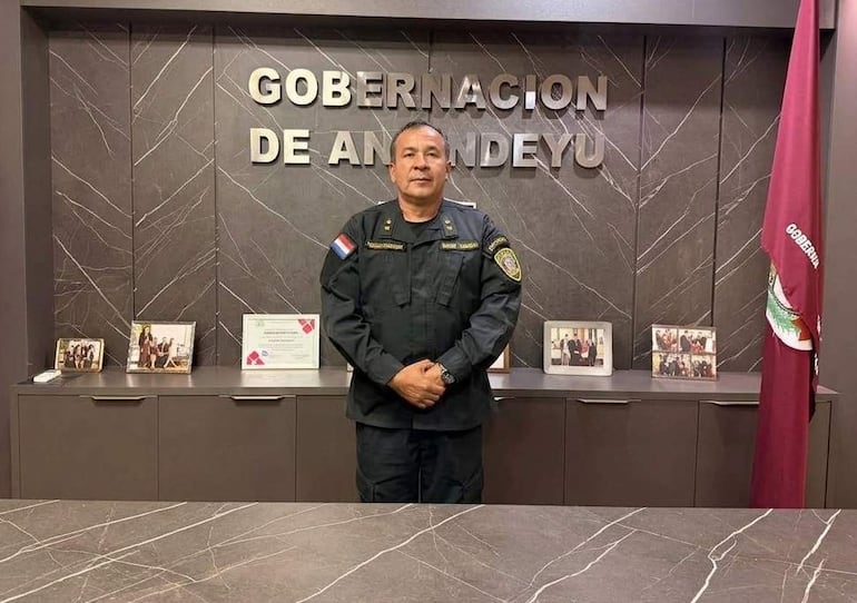 Ramón Escobar en uniforme oscuro, con bandera paraguaya en brazo, posa en su oficina con documentos y fotografías sobre la mesa.