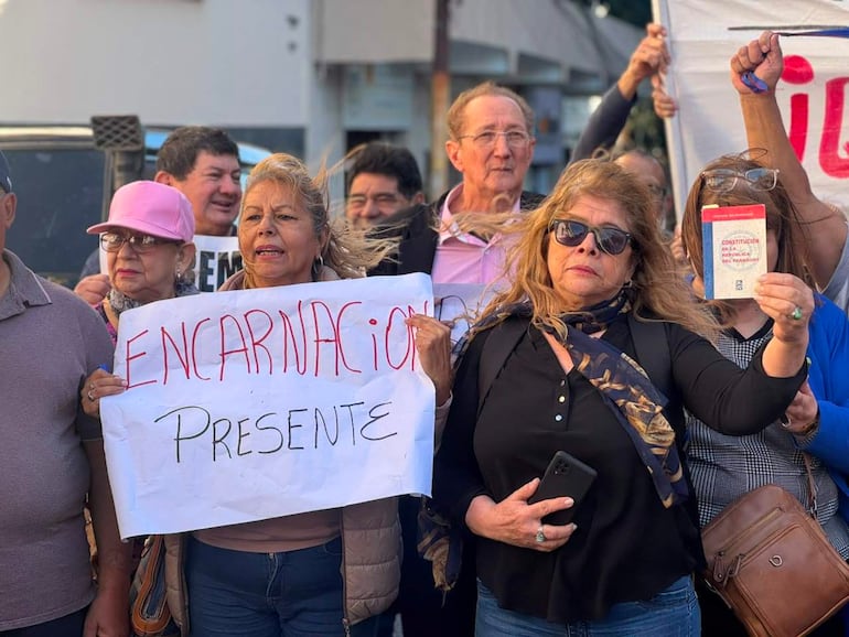 Mujer de cabello rizado sostiene un cartel en manifestación, acompañada de otra mujer con gorra y gafas. Ambiente de protesta en la ciudad.