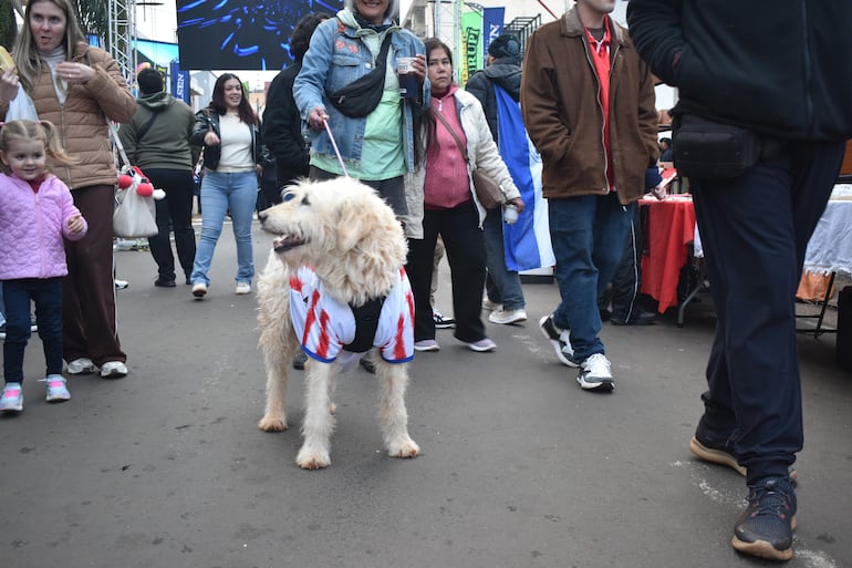 Mujer con snack junto a niña en abrigo rosa y perro con camiseta de la selección paraguaya, en un ambiente festivo.