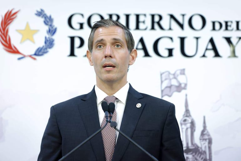 Marco Riquelme en traje oscuro y corbata, de pie frente a un micrófono con un cartel del Gobierno de Paraguay al fondo.