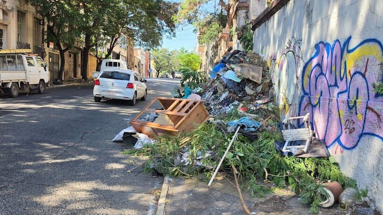 Gran bulto de basura en calle de Asunción, con muebles y plásticos dispersos, y graffiti colorido en la pared.